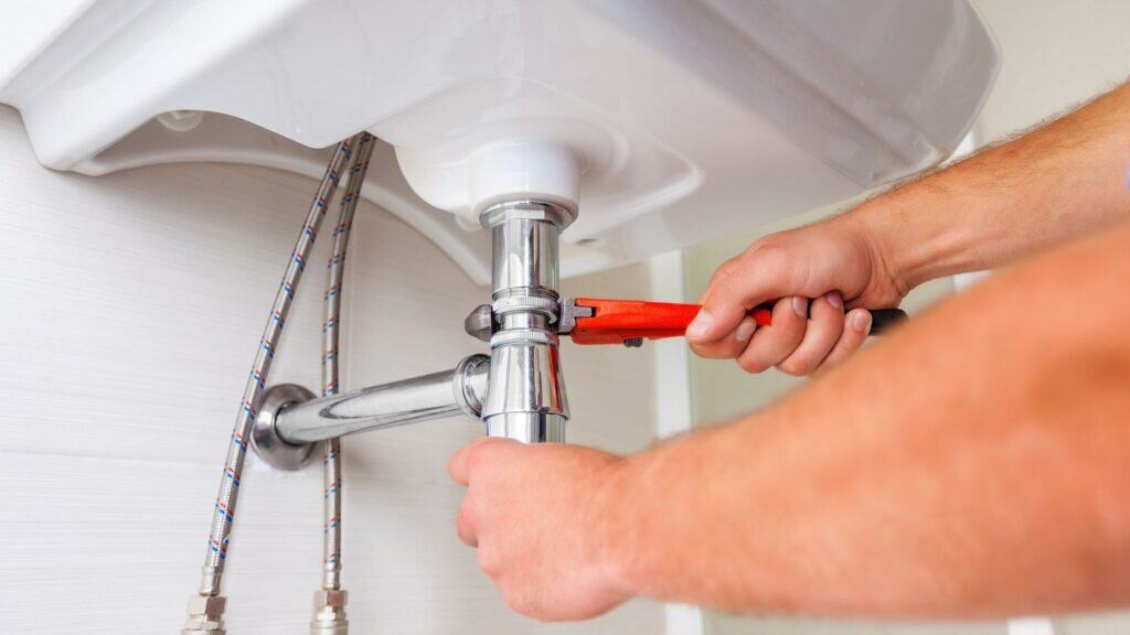 Hands holding a wrench, tightening a pipe under a white sink.