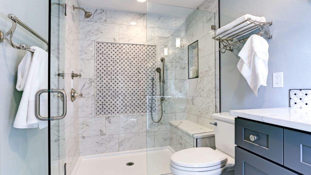 Modern bathroom with glass shower, marble tile walls, and a built-in shelf. A towel hangs on the rack, and a toilet is beside a dark-colored vanity with a white countertop.