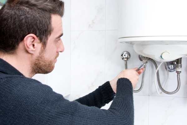 Person using a wrench to fix plumbing beneath a water heater attached to a tiled wall.