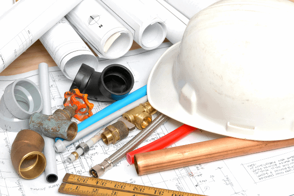 A collection of plumbing tools and materials, including pipes, a valve, fittings, a hard hat, and rolled blueprints, placed on a technical drawing sheet with a wooden ruler.