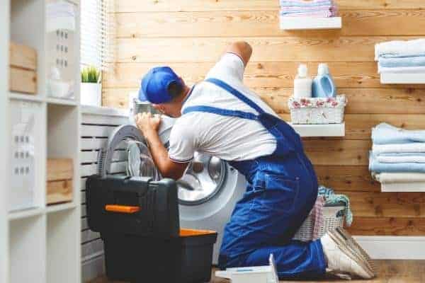 A maintenance worker in blue overalls and cap is repairing a front-load washing machine in a laundry room with wooden walls. A toolbox is on the floor nearby.