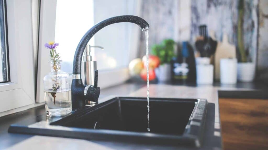 Kitchen sink with black faucet running water. A glass bottle with flowers and a soap dispenser are beside the sink. Sunlight illuminates the countertop.