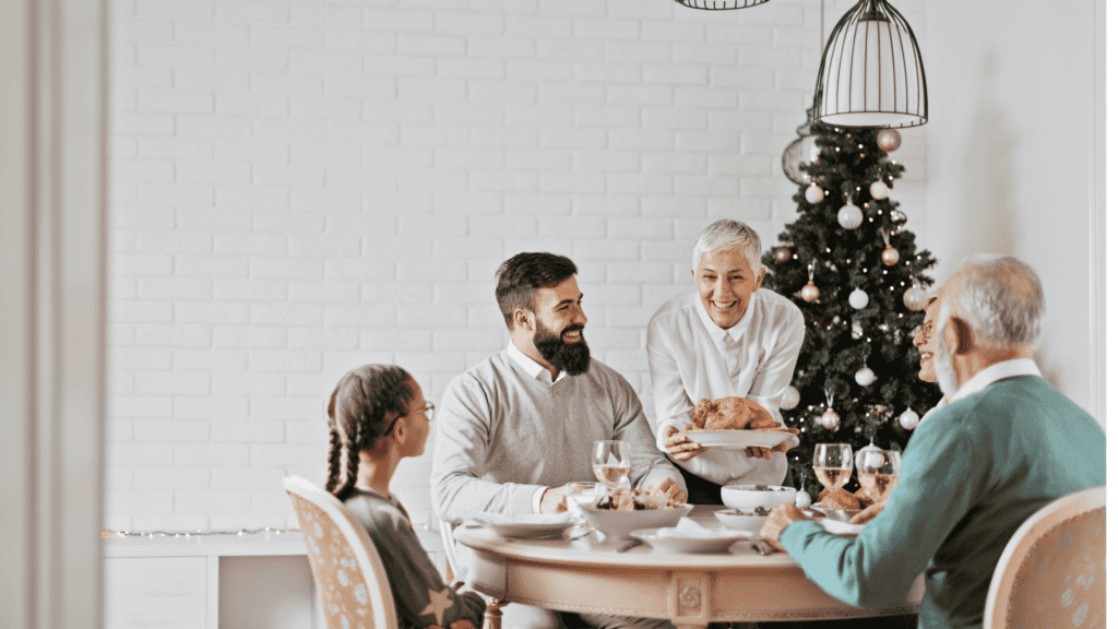 A family of four sits at a round table, sharing a meal. A woman serves a roast chicken. A decorated Christmas tree stands in the background.