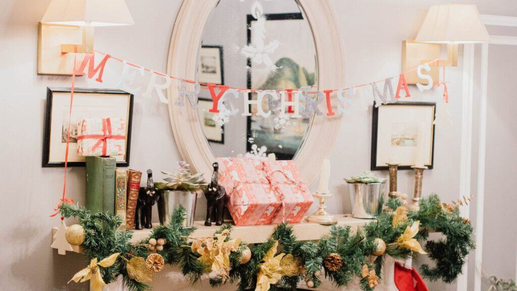 Festive mantel with Christmas garland, two wrapped gifts, books, and decorative reindeer figures. A "Merry Christmas" sign hangs above, and a mirror reflects some decoration.