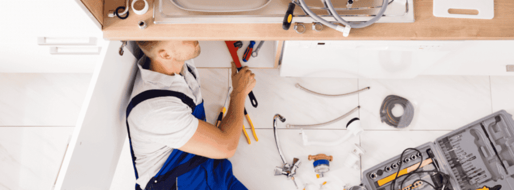 Person in overalls working under a kitchen sink, surrounded by plumbing tools and parts.