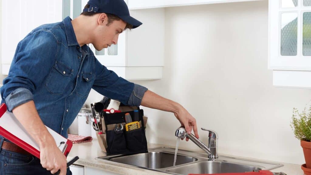 A person in a denim shirt and cap checks a running kitchen faucet. A tool bag is nearby on the counter.