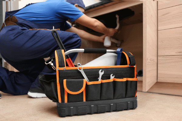 Person in blue overalls fixing pipes under a sink while crouched. A black and orange tool bag with various tools is in the foreground.