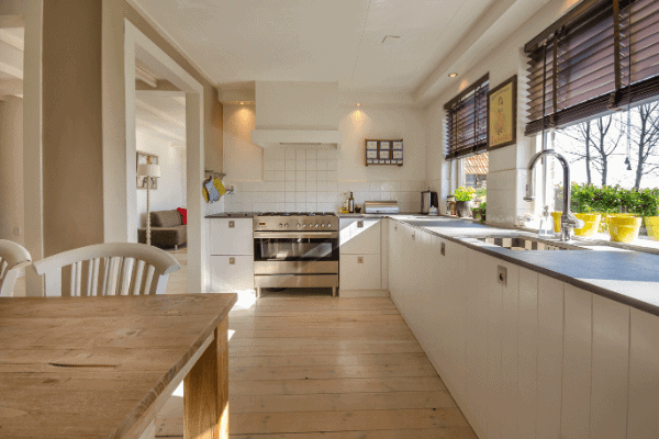 Spacious kitchen with wooden floors, white cabinets, and a stainless steel stove. A wooden table with white chairs is in the foreground. Large windows provide natural light.