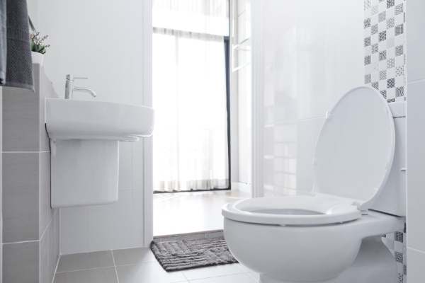 Modern bathroom with a white toilet and sink. Gray and white tiles decorate the walls. A window with sheer curtains allows natural light to enter. A gray mat is placed on the floor.
