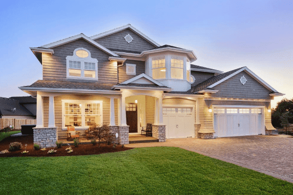 Two-story suburban house with stone and shingle exterior, illuminated at dusk. Features a large front porch, double garage, and manicured lawn.