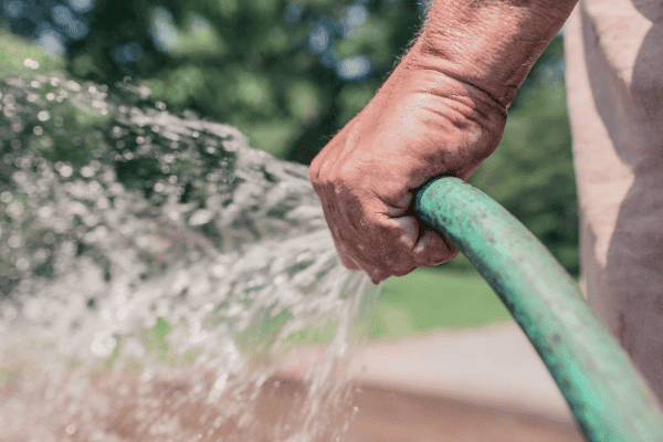A person using a green garden hose to water outdoors, with water visibly spraying from the nozzle.