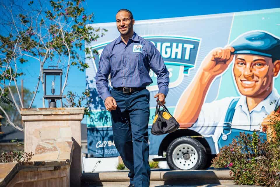 A man in a blue uniform walks outside, carrying a hat, with a colorful Plumbing Repair service van featuring a cartoon worker in the background.