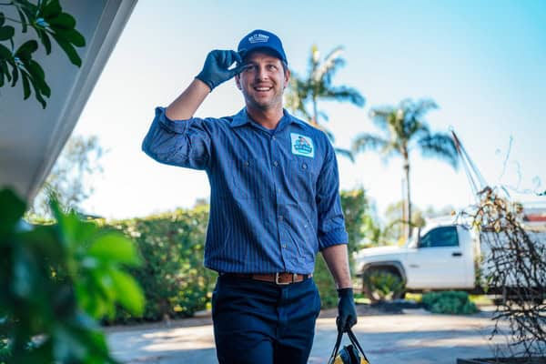 A man wearing a blue uniform and gloves smiles and tips his hat while standing outdoors near a white pickup truck and palm trees.