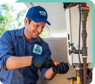 A technician in uniform and gloves uses a screwdriver to work on outdoor plumbing connected to a white appliance as part of a routine maintenance plan.