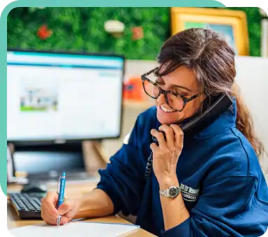 A woman wearing glasses discusses a maintenance plan over the phone while taking notes in her notebook at a desk, with a computer monitor in the background.